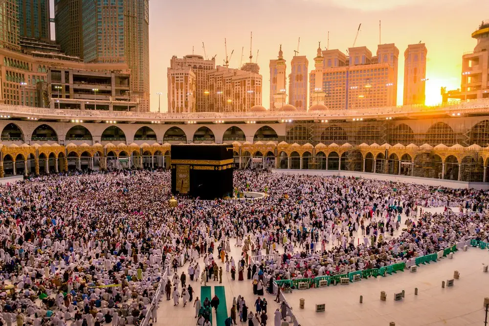 The Grand Mosque at sunset from above the courtyard, the Ka‘bah at the center of a dense crowd, the towers of the city catching the last light behind the arcade.