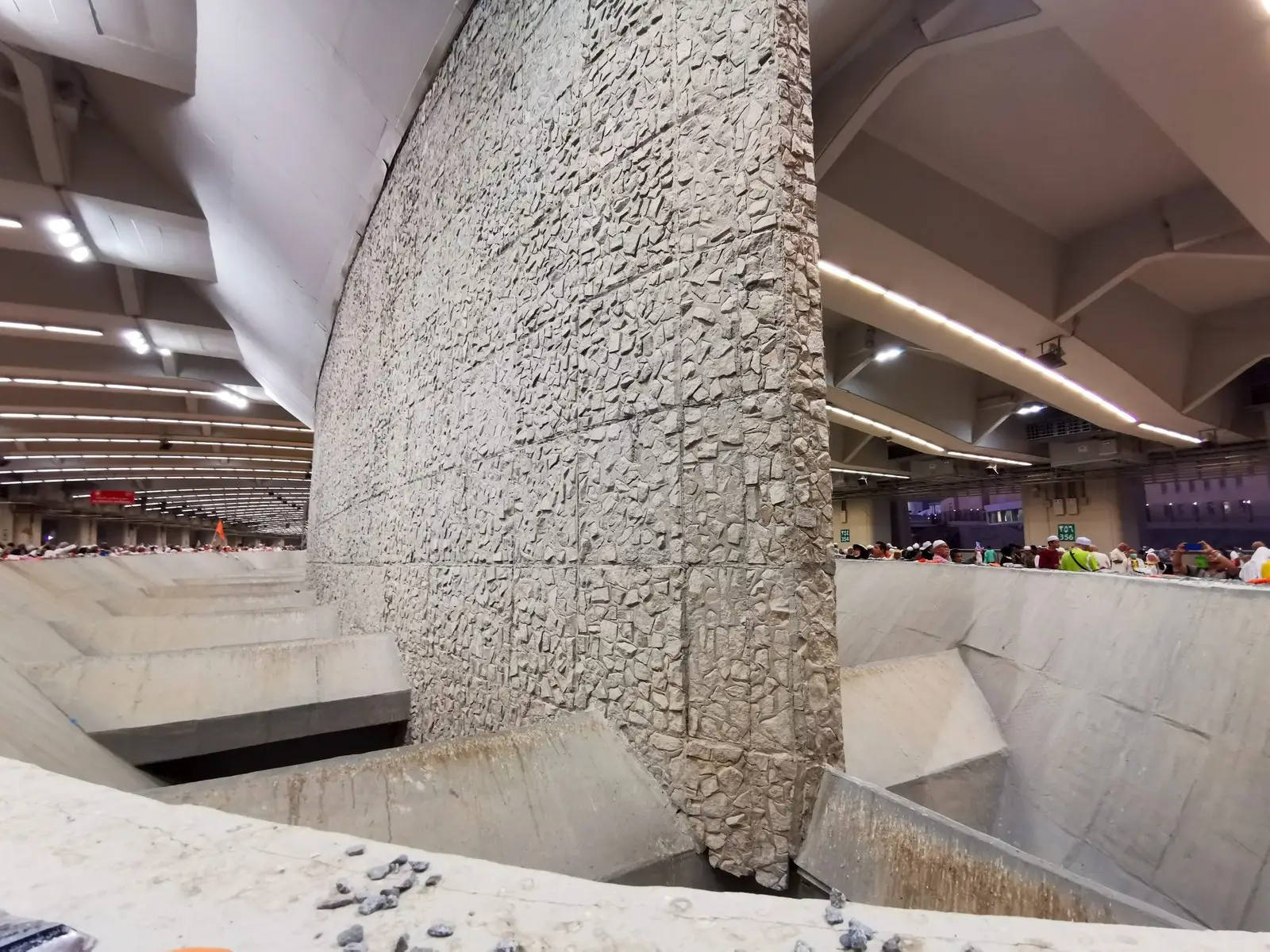 One of the Jamarat pillars inside the multi-story bridge at Mina: a tall wall of rough stone dropping into a concrete basin, with pilgrims at the rail on the level above.